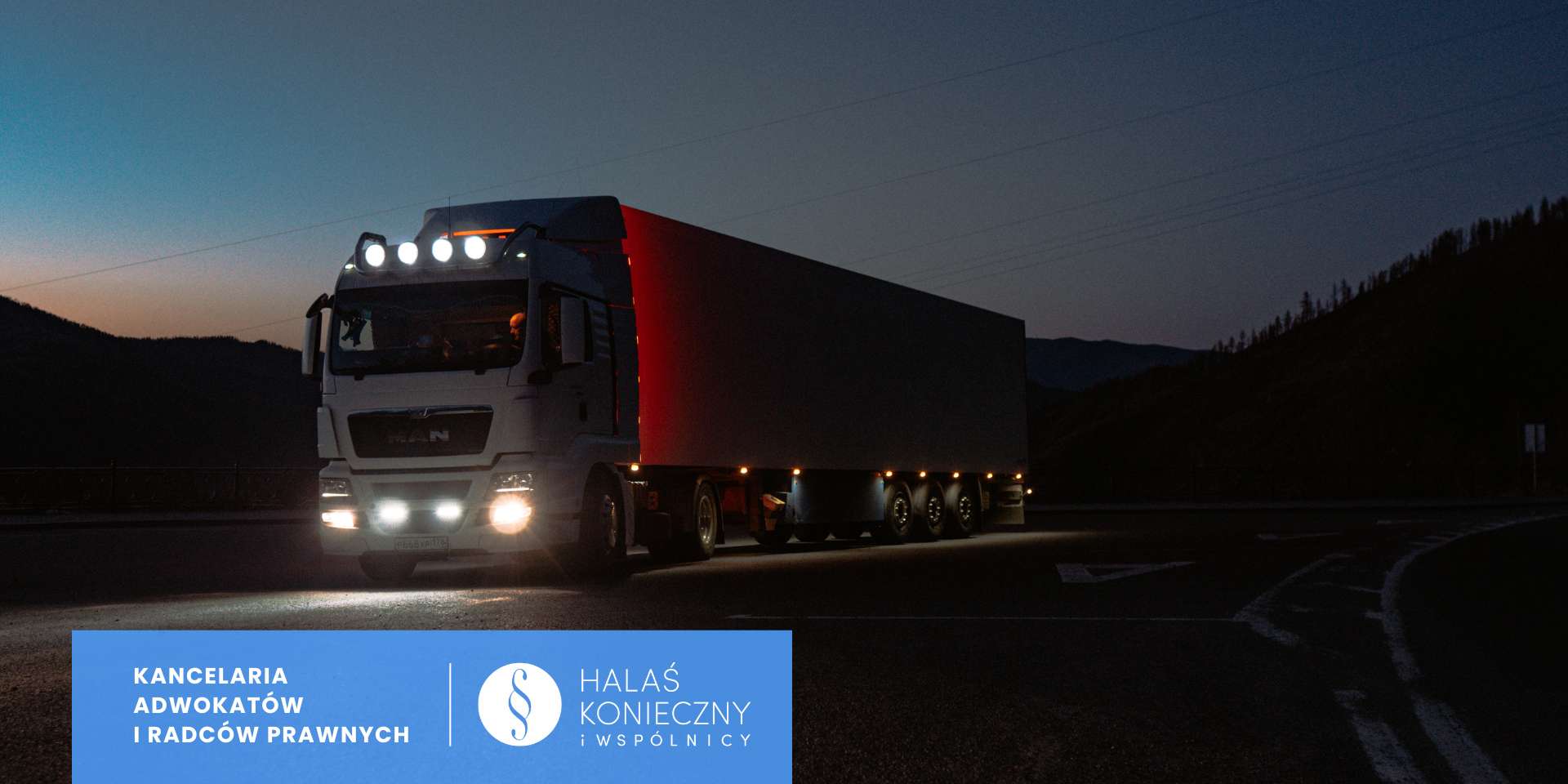 White semi-truck with illuminated headlights driving on a dark, winding road at dusk, mountains in the background, trailer trailing.