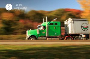Green semi-truck hauling a white trailer on a highway with autumn trees blurred in the background during motion