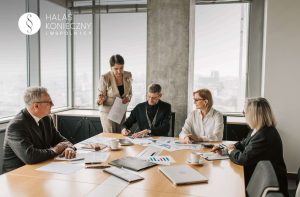 Group of professionals around a conference table reviewing documents; a woman stands presenting to the team.