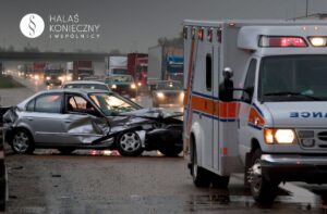 Car crash on a wet highway with damaged silver sedan and a nearby ambulance, emergency lights on, in rainy conditions.